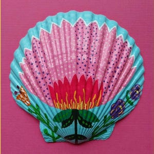 May include: A hand-painted seashell with a floral design. The shell is painted with a light blue border, a pink center with purple dots, and a red flower at the base. Green leaves and orange and purple accents complete the design.