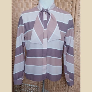 Puede incluir: Una blusa de manga larga a rayas con un gran cuello de lazo. La blusa es en tonos de rosa y morado.
