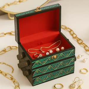 May include: A decorative wooden jewellery box with a red interior, featuring green and gold patterns. Inside, there's a gold necklace, ring, earrings, and pearls. Gold chains and other jewellery items are arranged around the box.