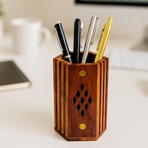 May include: A wooden pen holder with a geometric design, holding four pens. The holder is brown with a diamond-shaped cutout and brass accents. Pens are black, silver, and gold.
