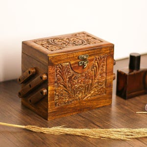 Peut inclure: Une boîte en bois avec des sculptures complexes de fleurs et de feuilles. La boîte a un couvercle à charnières avec un loquet en laiton et un petit trou de serrure. La boîte est en bois foncé et a un aspect rustique.