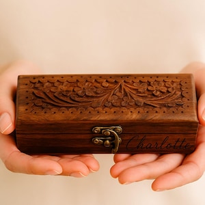 Könnte beinhalten: Eine rechteckige, dunkelbraune Holzbox mit aufwendigen Blumenschnitzereien auf dem Deckel. Die Box hat einen Metallverschluss und der Name "Charlotte" ist auf der Vorderseite eingraviert. Die Box wird in zwei Händen gehalten.