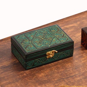Puede incluir: Una pequeña caja de madera tallada con un diseño verde y dorado. La caja tiene una tapa con bisagras y un cierre de latón.