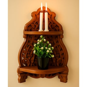 Może przedstawiać: Rzeźbiona drewniana półka narożna z dwiema zapalonymi świecami i małą rośliną doniczkową. Półka ma ciepły brązowy kolor i posiada skomplikowane rzeźbienia kwiatowe. Świece są białe, a roślina ma zielone liście i małe białe kwiaty.