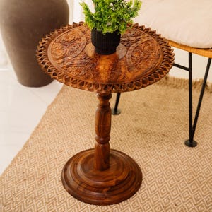 May include: A small, intricately carved wooden side table with a round top and a single pedestal base. The table is decorated with floral patterns and has a dark brown finish. A small potted plant sits on the table top.