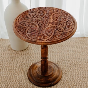 May include: A round wooden side table with intricate floral carvings. The table has a single pedestal base and a dark brown finish.