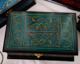 May include: Several decorative wooden boxes with intricate, hand-painted designs. The boxes are black with teal and gold accents, and feature ornate patterns. The boxes have gold-colored clasps and are arranged on a white surface.