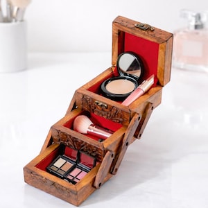 May include: A wooden makeup organizer with three pull-out drawers, each lined with red fabric. The top drawer contains a compact mirror and a powder compact. The middle drawer holds a makeup brush, and the bottom drawer has eyeshadow palettes.
