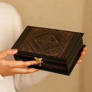 Peut inclure: Une boîte en bois noire avec des motifs floraux et géométriques dorés complexes sur le couvercle. La boîte a un fermoir doré et est tenue par une personne portant une chemise blanche. C'est un objet de rangement décoratif.