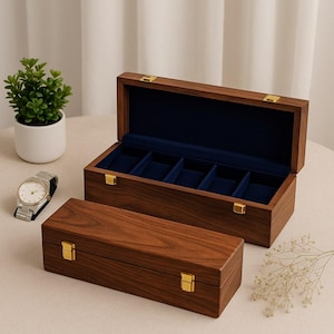 Op de afbeelding: Twee houten horlogeboxen, één open en één gesloten, met goudkleurige sluitingen. De open box onthult een blauw fluwelen interieur met vijf compartimenten. Een zilveren polshorloge met een witte wijzerplaat staat in de buurt. De dozen zijn gemaakt van donker hout.