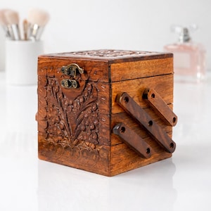 May include: A wooden jewelry box with intricate floral carvings and a hinged, tiered design. The box is a warm brown color, with a metal clasp and hinges. Makeup brushes and a perfume bottle are in the background.