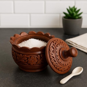 May include: A wooden salt cellar with a carved floral design, filled with white salt. The lid is partially open, revealing the contents. A small white spoon rests beside the container. The salt cellar is brown and sits on a dark surface.