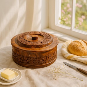 May include: A round, wooden butter dish with a lid featuring intricate floral carvings. The dish is brown and sits on a table next to a small plate with a pat of butter, a loaf of bread, and a butter knife.