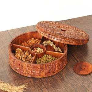 Könnte beinhalten: Eine runde Holzkiste mit einem geschnitzten Deckel, unterteilt in fünf Fächer, gefüllt mit verschiedenen Nüssen und Trockenfrüchten. Die Kiste ist aus dunkelbraunem Holz und hat aufwendige Schnitzereien.
