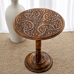 May include: A round, brown wooden side table with intricate floral carvings on the tabletop. The table has a single pedestal base and a circular foot. The table is approximately 24 inches in height and 20 inches in diameter.