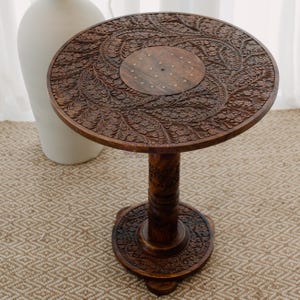 May include: A round, intricately carved wooden side table with a floral design. The table has a single, thick, carved pedestal base.