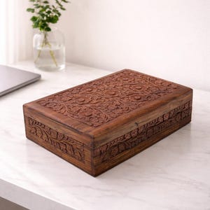 May include: A rectangular, dark brown wooden box with intricate floral carvings on the lid and sides. The box is placed on a white marble surface, with a glass vase of greenery in the background. The box is a decorative storage item.