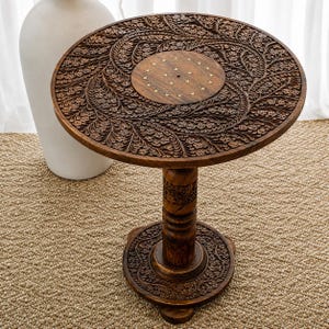 May include: A dark brown, intricately carved wooden side table. The round tabletop features a floral design and a central circle with brass accents. The table has a carved pedestal base and three feet. The table is approximately 24 inches tall.