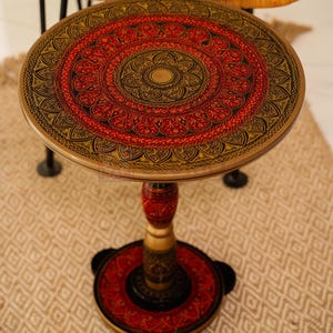 May include: A round wooden side table with a red and gold mandala design on the top. The table has a single pedestal base with a red and gold design.
