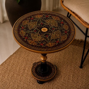 May include: A round, decorative side table with a dark, patterned top featuring floral and geometric designs in red, gold, and green. The table has a gold-colored rim and a pedestal base. The table is approximately 20 inches in diameter.