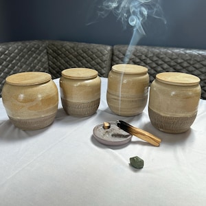 Puede incluir: Cuatro tarros de cerámica redondos con tapa, cada uno con un acabado bicolor en beige y marrón texturizado. Una pequeña bandeja circular contiene un trozo de madera de palo santo encendido que emite humo, junto a una pequeña piedra verde.