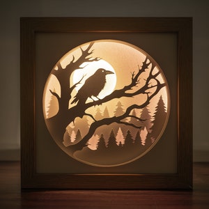 May include: A wooden framed shadow box with a lit up paper cut design of a raven perched on a branch in a forest. The raven is black and the forest is silhouetted against a glowing moon.