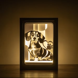 Könnte beinhalten: Eine schwarze und weiße Silhouette eines Dackels mit einem Pfotenabdruck in einem weißen Rahmen, durch den ein warmes weißes Licht scheint.
