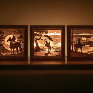 May include: Three wooden shadow boxes with illuminated silhouettes of cowboys riding horses in a desert landscape. Each box features a different scene with a moon, trees, and mountains.