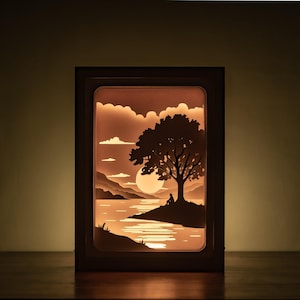 Op de afbeelding: Een verlichte papieren lichtbak in een houten frame, met een serene landschapsscène. De afbeelding toont een boom, een ondergaande zon en een persoon zittend op een eiland. Het warme kleurenpalet, met oranje en bruine tinten, creëert een rustige sfeer.