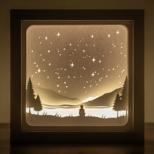 May include: A square shadow box frame containing a layered paper cutout scene. The backlit display features a silhouette of a person sitting by a lake, surrounded by pine trees and mountains under a night sky filled with numerous cut-out stars.