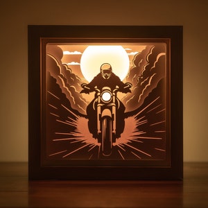 May include: A 3D paper cut-out lamp with a silhouette of a person riding a motorcycle through a cloud-filled sky. The lamp is lit from behind, creating a warm glow.