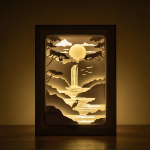 Op de afbeelding: Een wit houten frame met een uitgesneden ontwerp van een waterval, maan, bomen en een vogel in een meer. Het frame wordt van achteren verlicht, waardoor een gloeiend effect ontstaat.