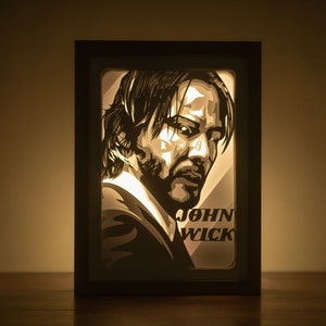 May include: A black framed light-up wall art piece featuring a silhouette of John Wick, a fictional character from the John Wick film series. The text "John Wick" is visible below the silhouette.