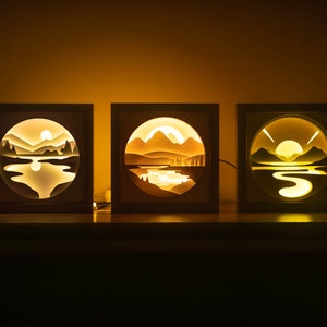 Peut inclure: Trois boîtes à lumière en bois avec des scènes illuminées de montagnes, de lacs et de couchers de soleil. Chaque boîte présente une scène différente.