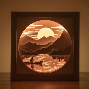May include: A wooden shadow box with a circular cutout featuring a paper cut-out design of a mountain lake scene with a person fishing in a boat at sunset.