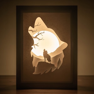 May include: A wooden frame with a white paper cutout of a wolf howling at the moon. The wolf is silhouetted against a full moon and a tree. The frame is lit from behind, creating a glowing effect.