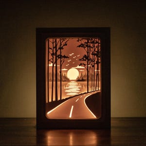 Op de afbeelding: Een ingelijst, verlicht papieren kunstwerk dat een weg afbeeldt die naar een zonsondergang boven een meer leidt. Het kunstwerk toont silhouetten van bomen, vogels en een warme, oranje gloed, waardoor een serene landschapsbeeld ontstaat.