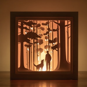 May include: A wooden shadow box with a light inside, depicting a silhouette of a person walking a dog through a forest. The scene is cut out of paper and illuminated from behind.