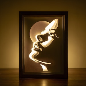 Può includere: Un'opera d'arte in carta incorniciata con una silhouette di un viso di donna. L'opera è illuminata da dietro, creando un bagliore morbido.