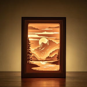 以下が含まれることがあります： 満月のある山岳風景の3Dペーパーカットデザインが施された木製の影箱。シーンは後ろから照らされ、温かい光を放っています。