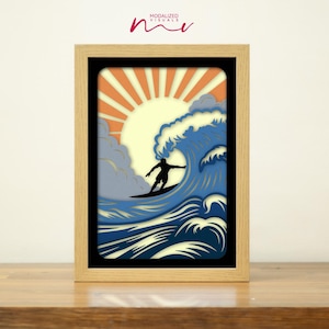 May include: A framed art print featuring a surfer riding a large wave under a setting sun. The artwork uses layered paper to create a 3D effect, with shades of blue, orange, and yellow. The frame is made of light-colored wood. The text "MODALIZED VISUALS" is in the upper left corner.