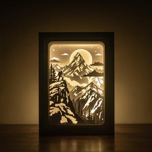 May include: A framed paper art piece showing a mountain landscape with a hiker, trees, and a full moon. The artwork is lit from within, producing a warm glow. The frame is dark, and the theme is nature and adventure. The dimensions are in centimeters.