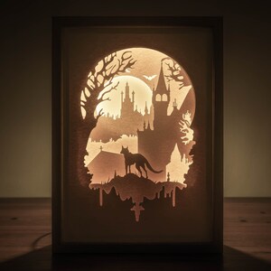 May include: A light-up shadow box featuring a paper cut-out design of a spooky castle, a wolf, and a bat. The castle is illuminated from behind, creating a glowing effect.