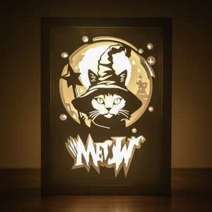 May include: A black cat wearing a witch's hat and holding a wand, with a full moon behind it. The word "MEOW" is written below the cat in a stylized font. The image is illuminated from behind, creating a glow effect.