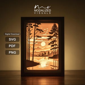 Op de afbeelding: Een rechthoekige schaduwdooslijst met een gelaagd papier-gesneden landschap van een meer, dennenbomen en bergen bij zonsondergang, van binnenuit verlicht. De tekst bovenaan luidt MODALIZED VISUALS, links staat Digital Download, SVG, PDF, PNG.