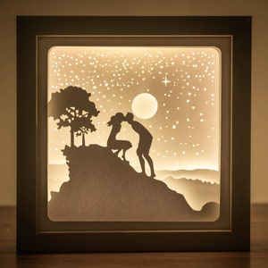 Può includere: Una scatola di luce bianca con una silhouette di una coppia che si bacia su una collina sotto un cielo stellato. La coppia è in silhouette contro una luna piena.