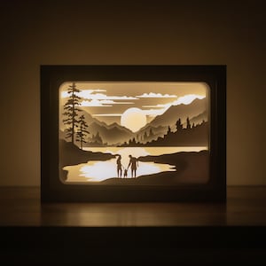 May include: A framed, layered paper art piece depicting a family holding hands by a lake at sunset. The artwork features silhouettes of trees, mountains, and a large sun. The frame is dark brown, and the scene is illuminated from within, creating a warm glow.