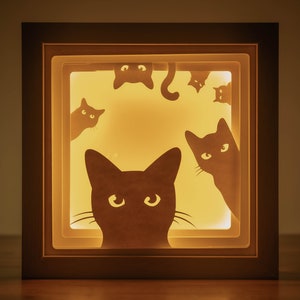 May include: A wooden shadow box with a light inside, featuring a silhouette of five black cats looking out from the box. The cats are cut out of paper and are arranged in a playful, whimsical design.
