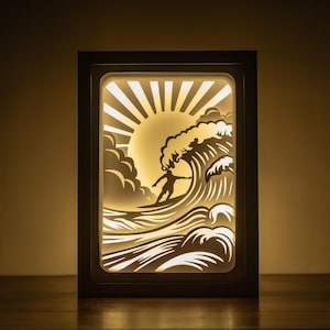 May include: A white wooden frame with a light-up design of a surfer riding a wave with a sun rising in the background.