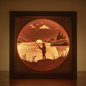 May include: A wooden shadow box with a paper cut-out design of a person fishing at sunset. The scene includes a lake, trees, and a setting sun.